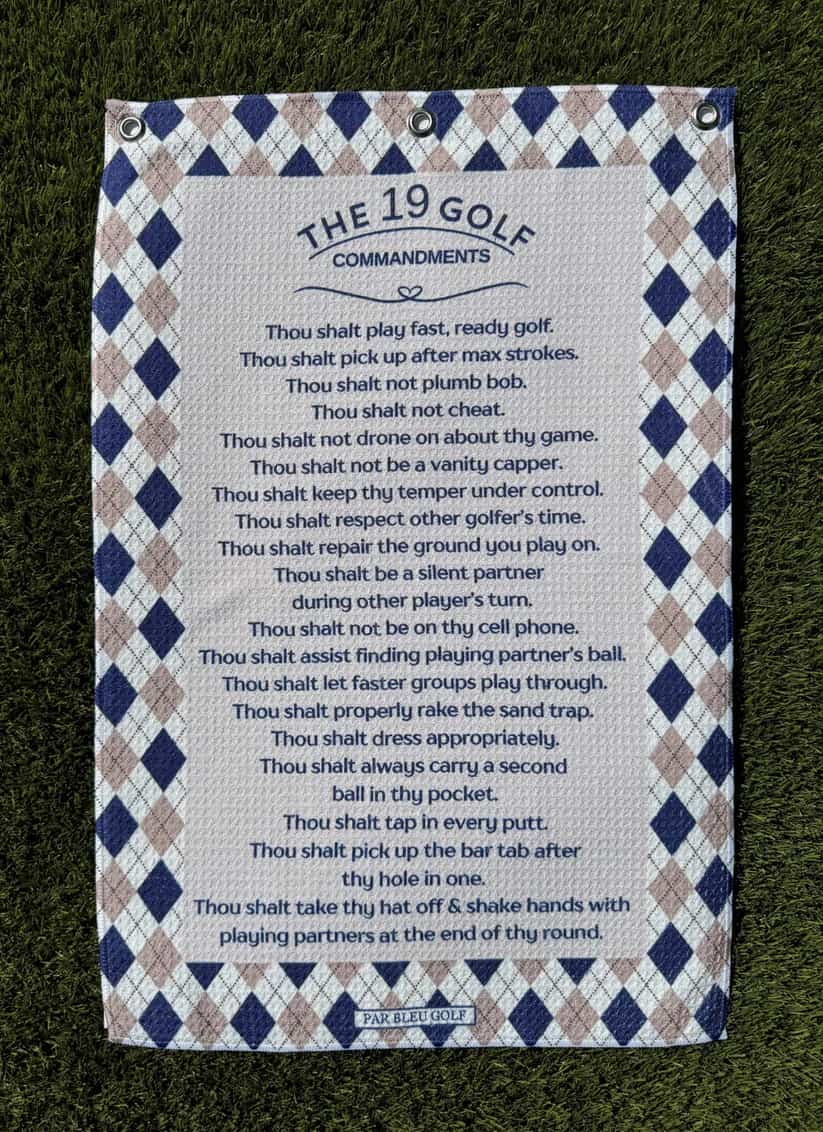 The 19 Golf Commandments towel laid flat on grass showing full printed list and argyle frame