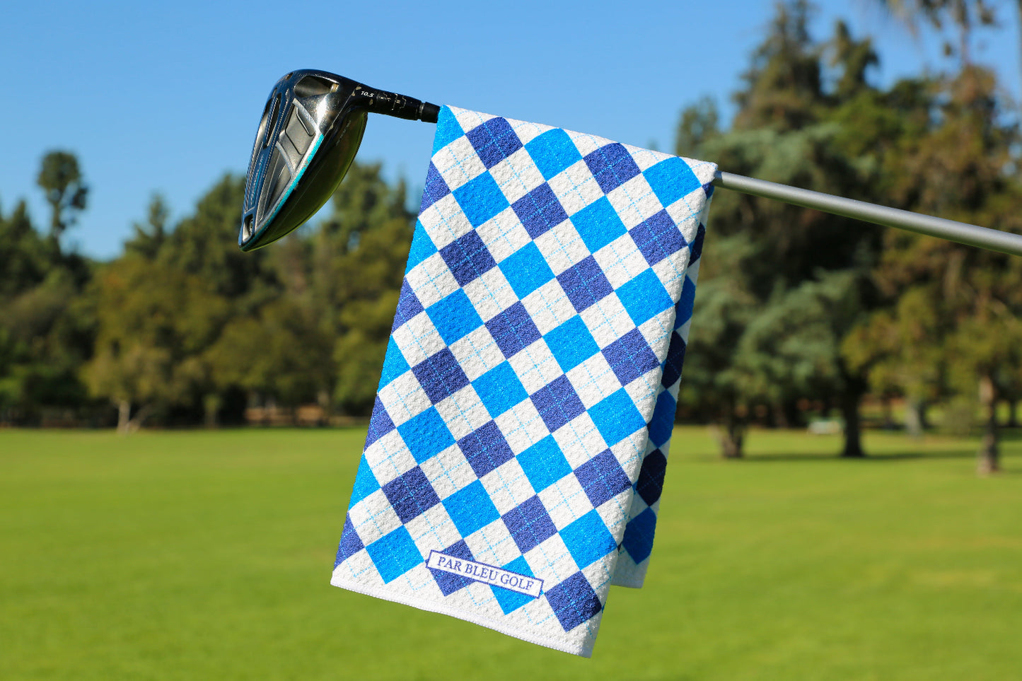 Blue argyle golf towel hanging on club with green fairway and trees in background