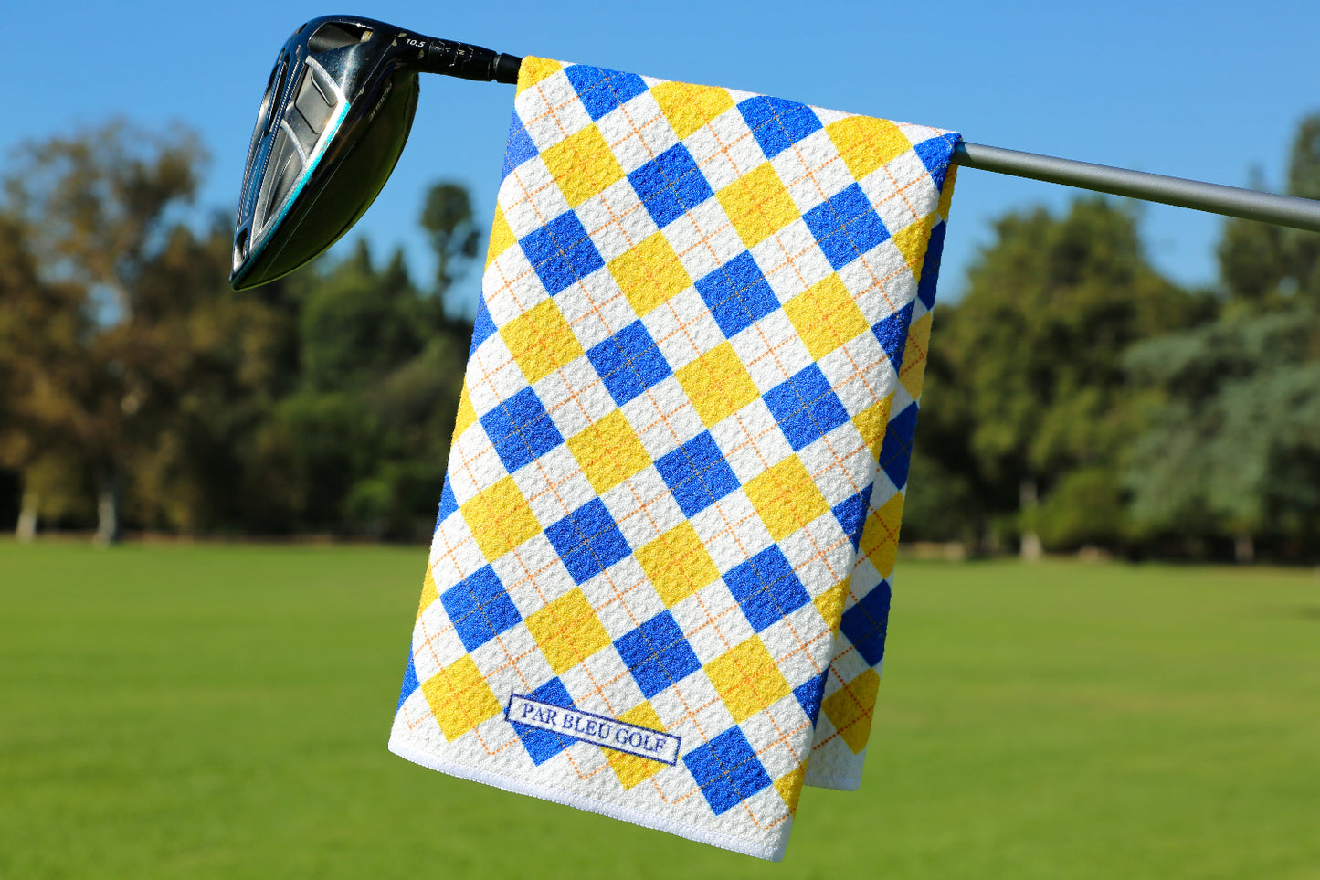 Blue and gold argyle golf towel hanging on club over green fairway with trees and blue sky