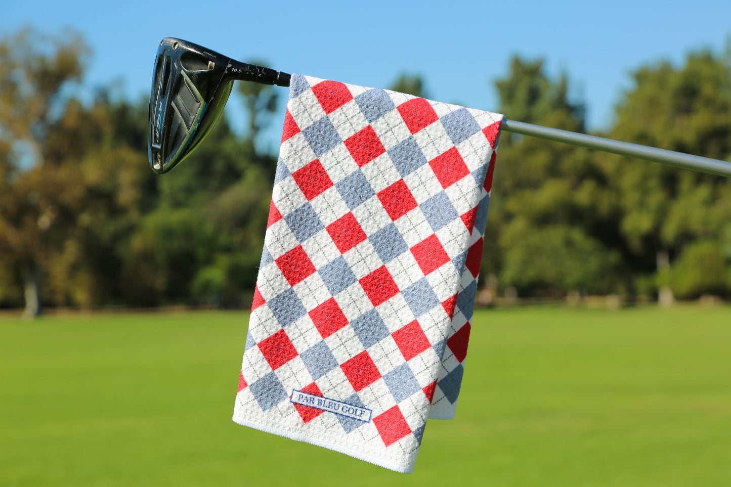 Red and grey argyle golf towel hanging on club with green fairway and blue sky in background