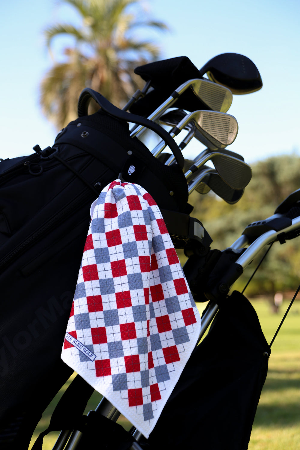 Red and grey argyle golf towel clipped to black golf bag on sunny course