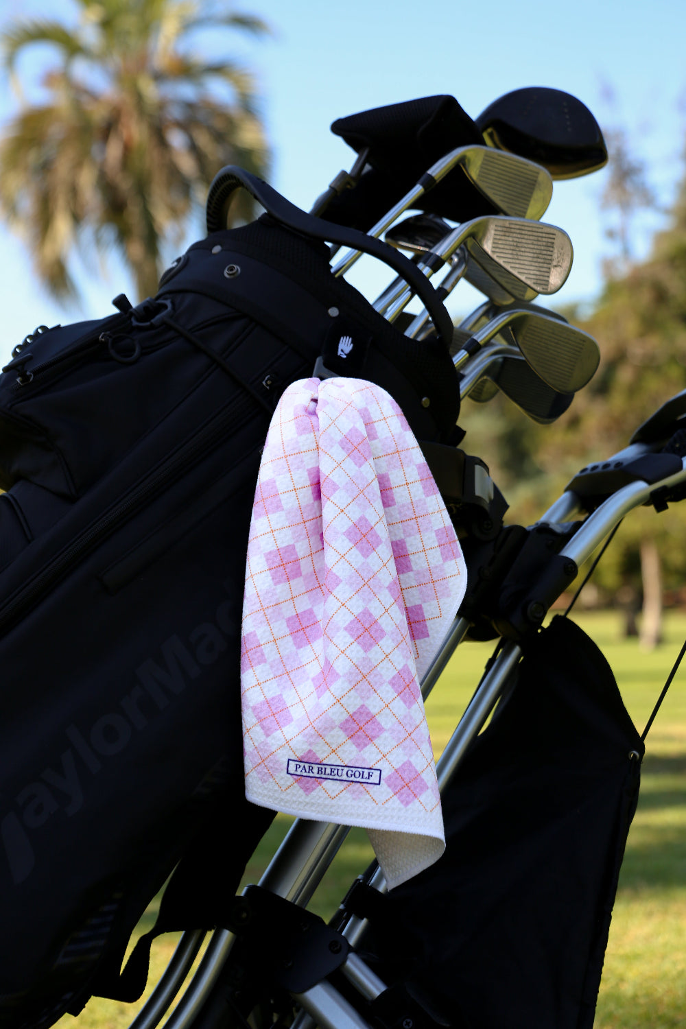 Light pink argyle golf towel clipped to black golf bag with palm trees in the background