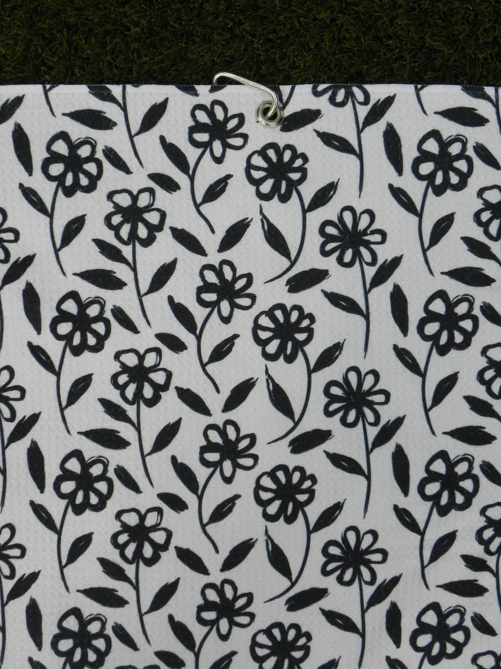 Golf towel with black floral daisy print hanging by carabiner clip on grass