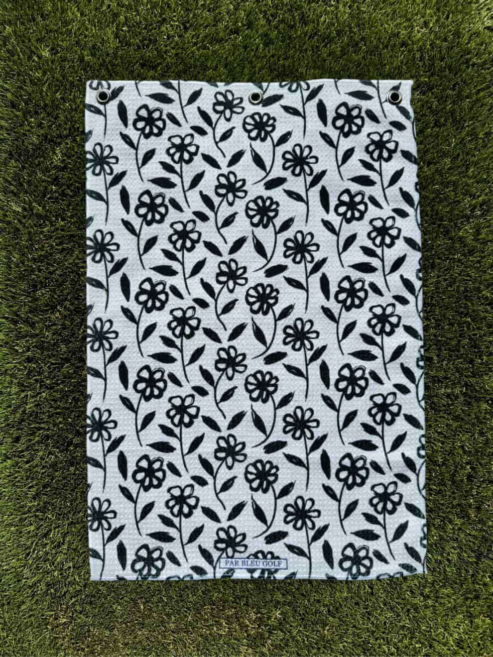 Black daisy floral golf towel laid flat on grass, showing full pattern and soft microfiber material