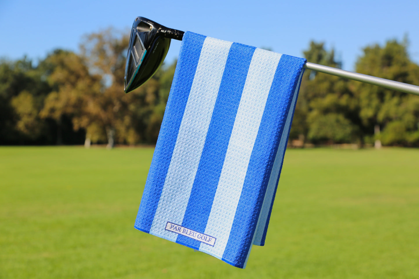 Blue and navy striped golf towel hanging on a club over green grass and trees