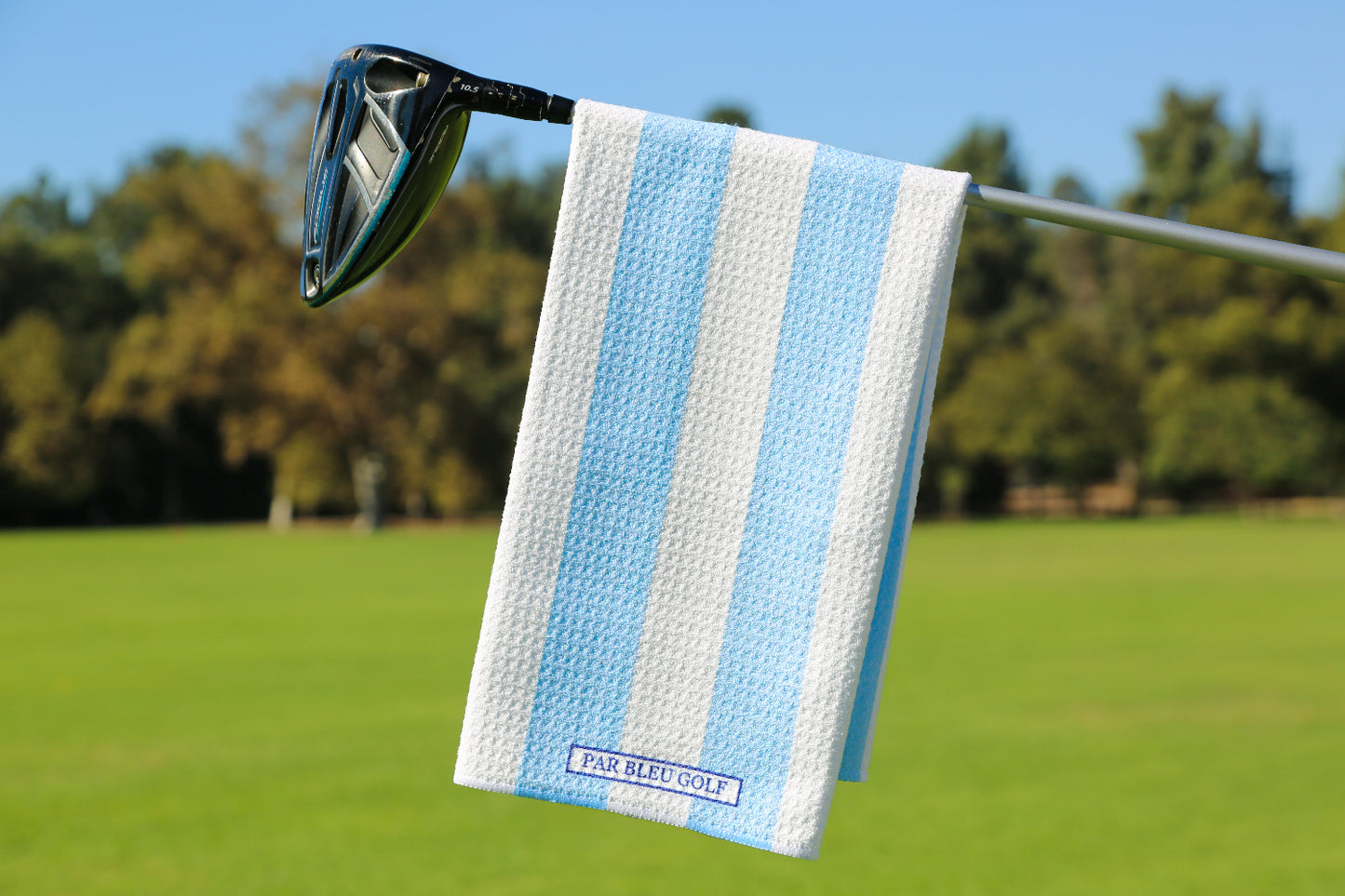 Blue and white striped golf towel hanging on club with trees and clear sky in background