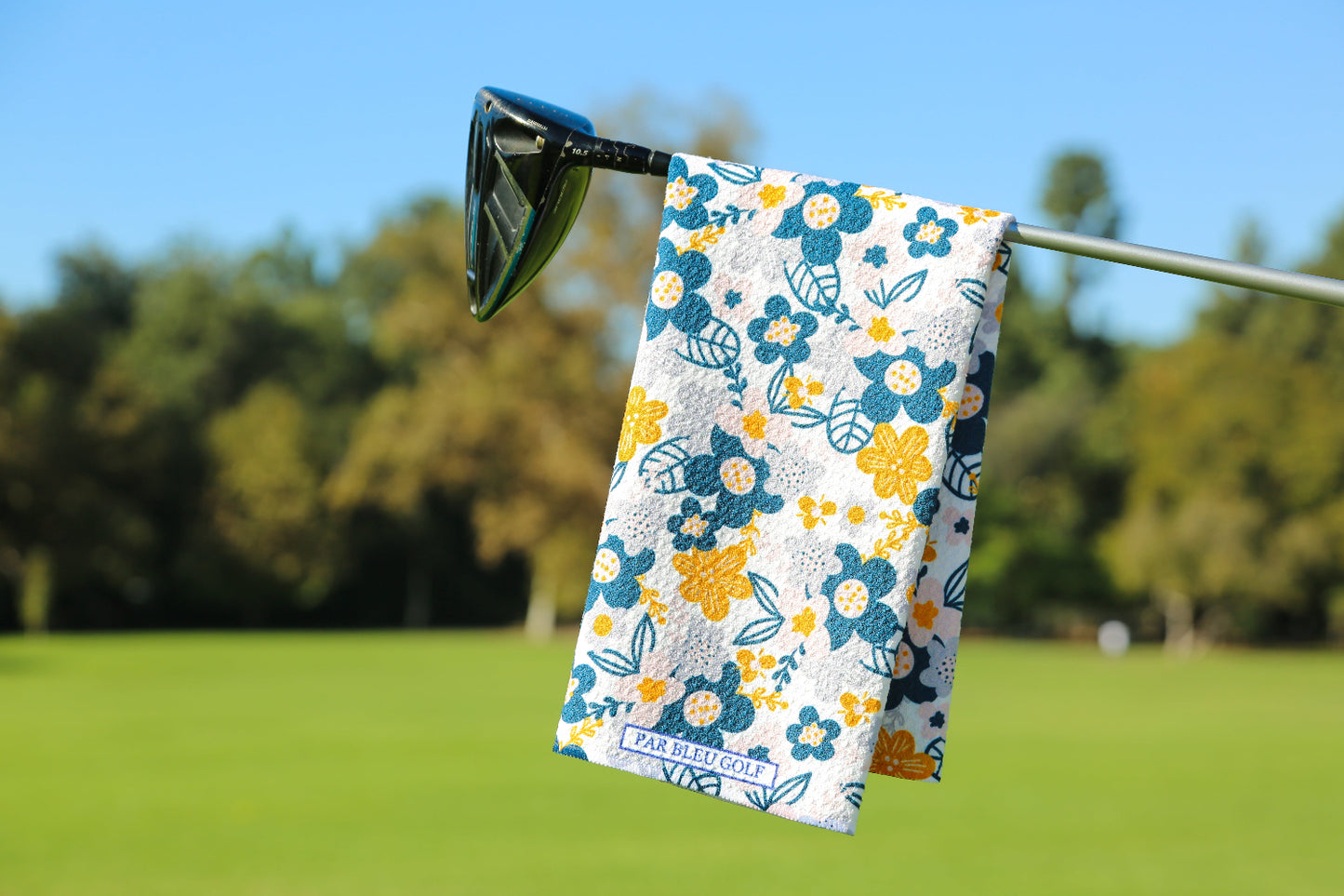 Stylish women’s golf towel with grey and yellow floral design hanging on a club against green fairway