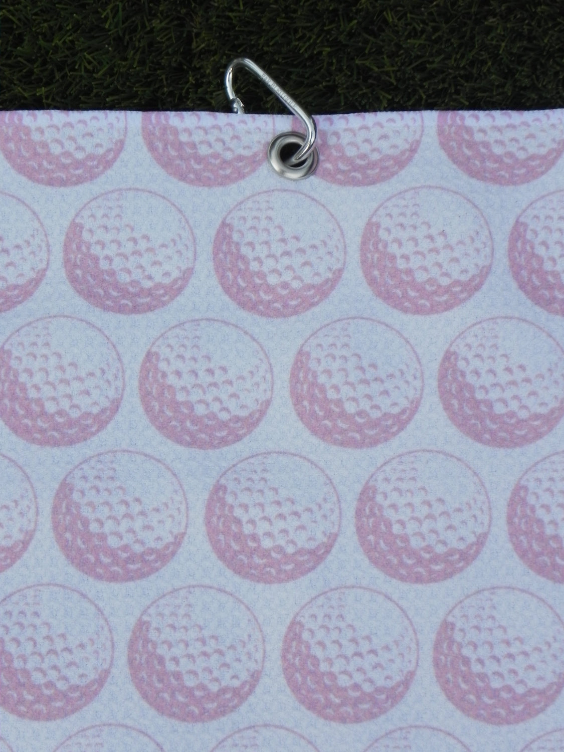 Pink golf towel hanging by metal carabiner, showing repeating golf ball print