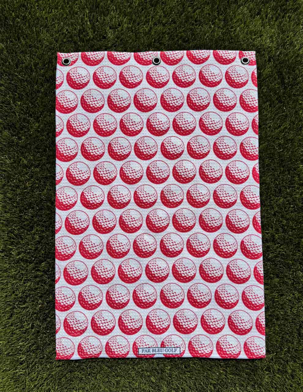 Red microfiber golf towel with golf ball print laid flat on green grass