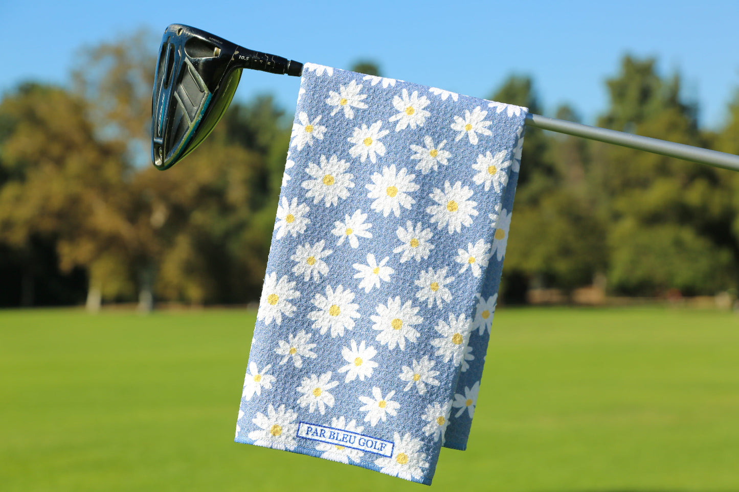 Grey golf towel with white daisy pattern hanging on club over fairway with trees and blue sky
