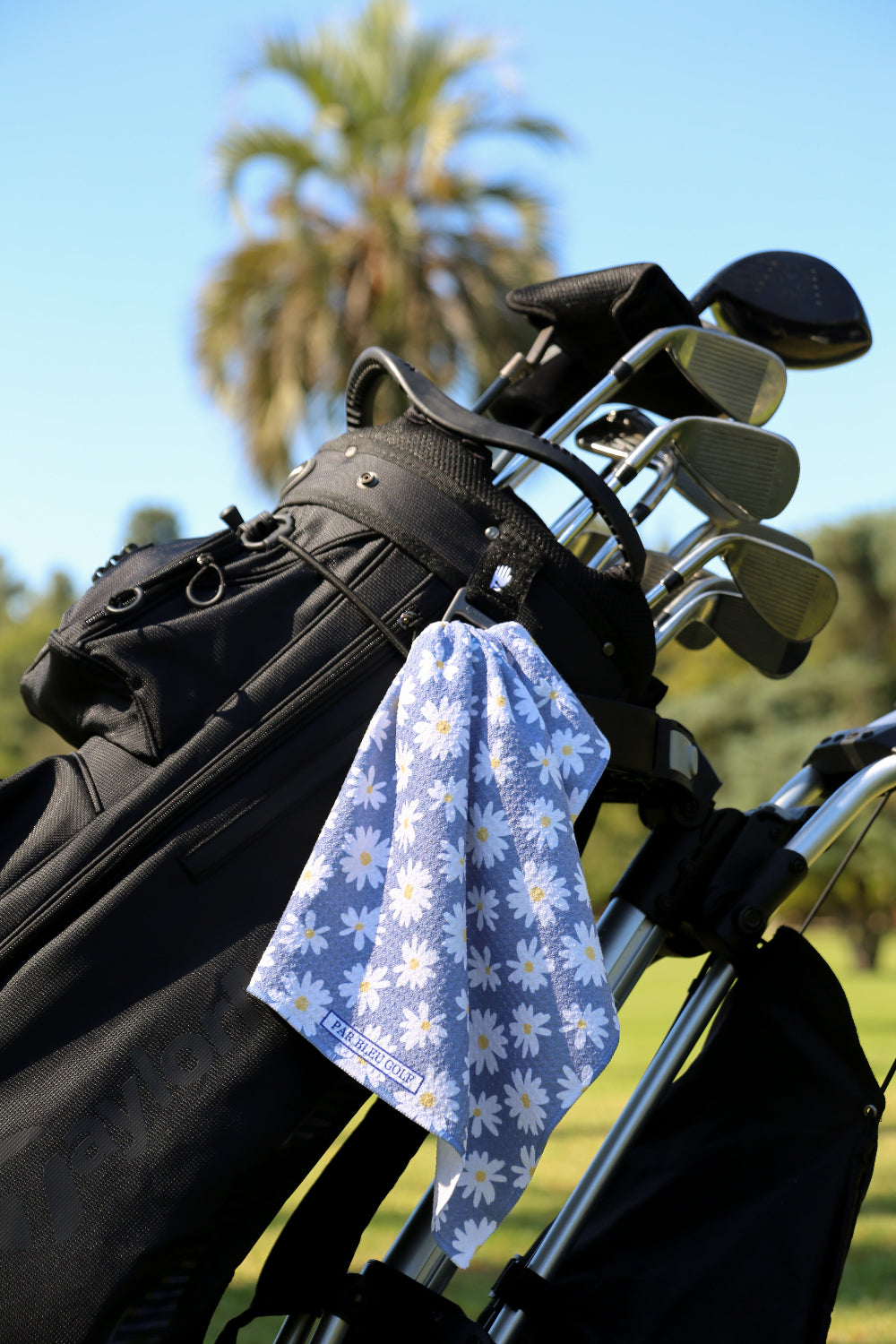 Grey daisy print golf towel clipped to black golf bag with palm trees in background