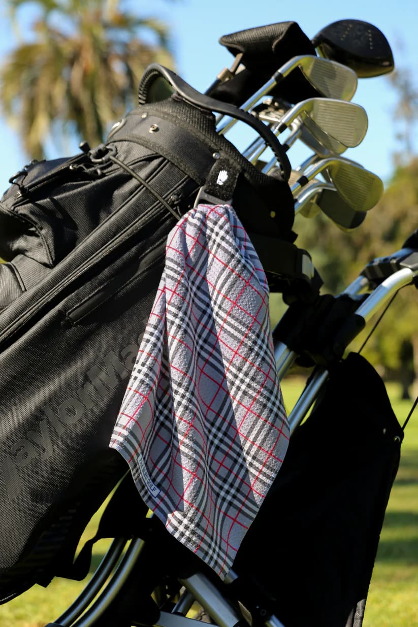 Grey plaid golf towel clipped to a black golf bag with palm trees and blue sky in the background