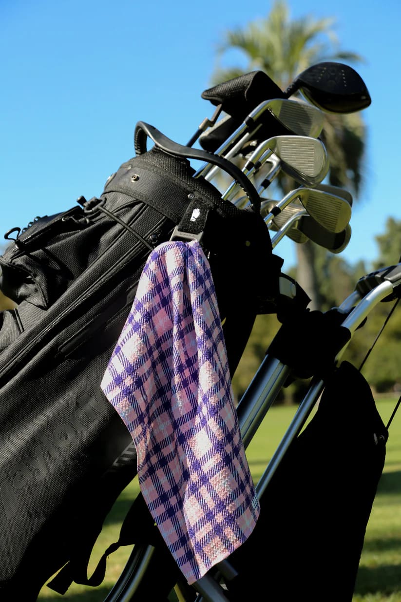 Pink plaid golf towel clipped to black golf bag on sunny day with palm trees in background