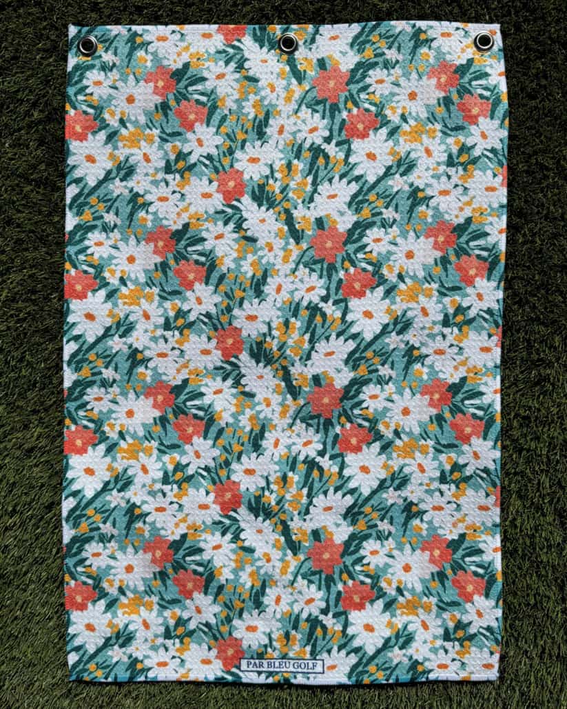 Women’s golf towel laid flat on grass, showcasing teal and orange daisy print and lightweight microfiber fabric