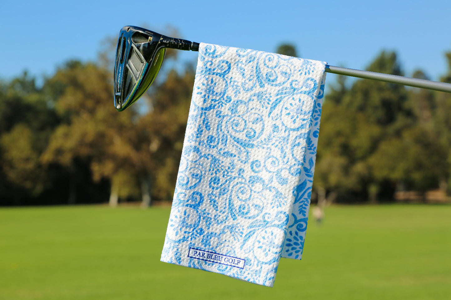 The Toile: Blue microfiber golf towel with floral pattern hanging on club over golf course