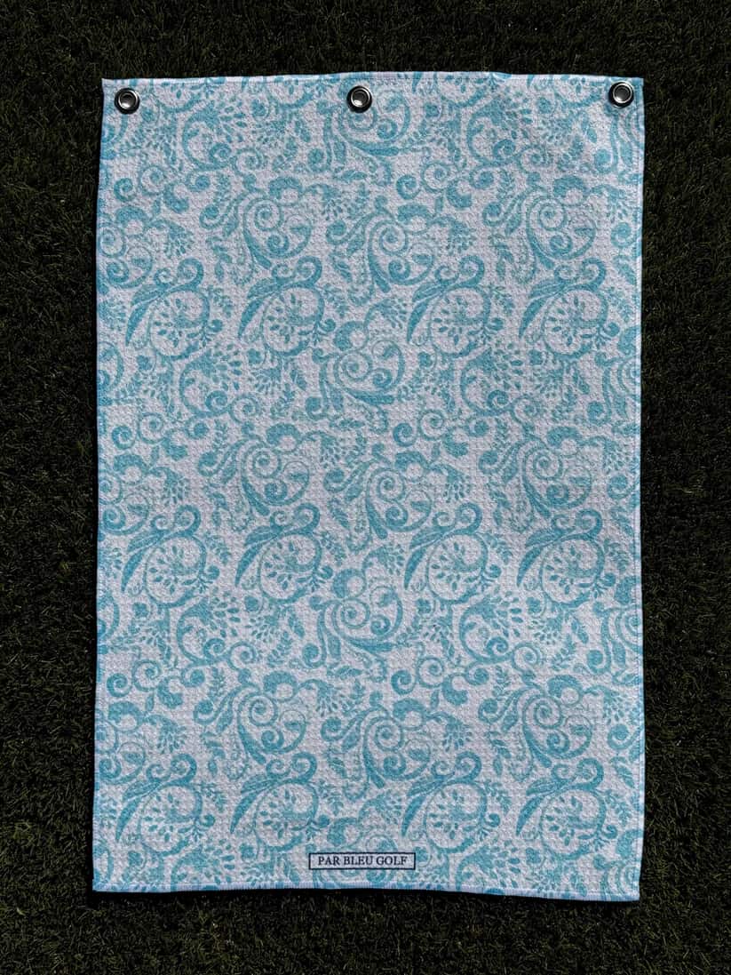 Flatlay of The Toile: Teal golf towel on green grass, featuring delicate damask-style design
