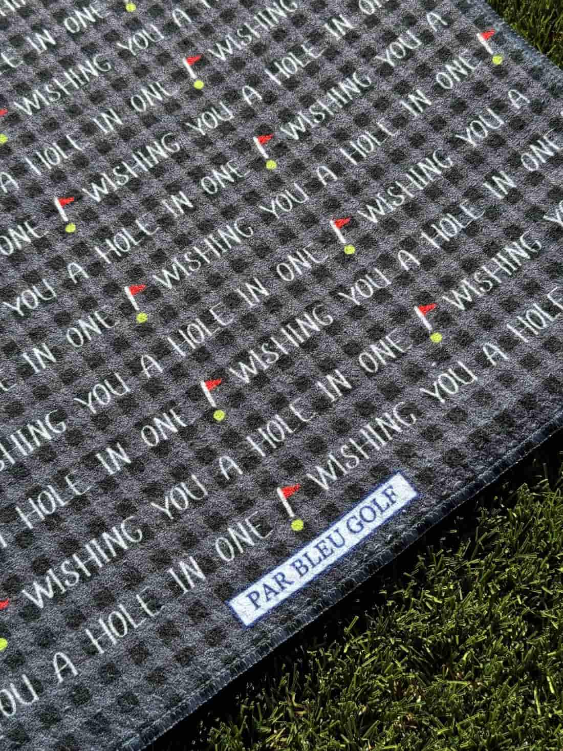 Close-up of Par Bleu black golf towel with “Wishing You a Hole in One” text and stitched brand tag
