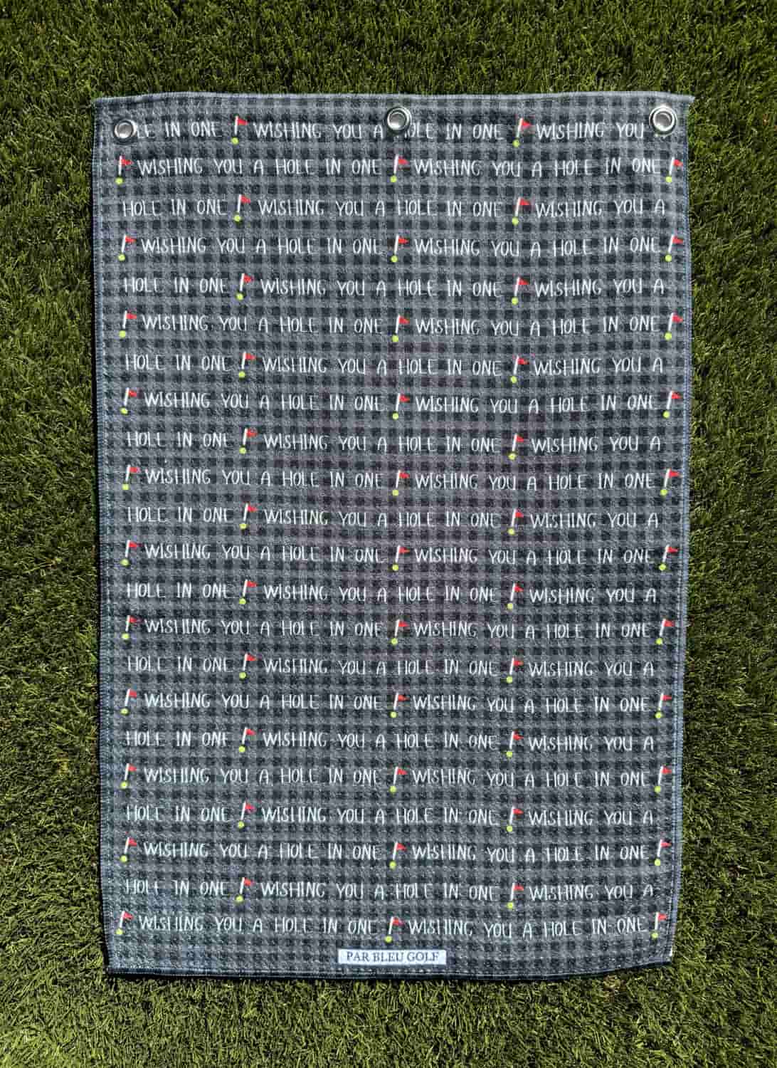 Flat black microfiber golf towel featuring repeating “Wishing You a Hole in One” print, shown on grass