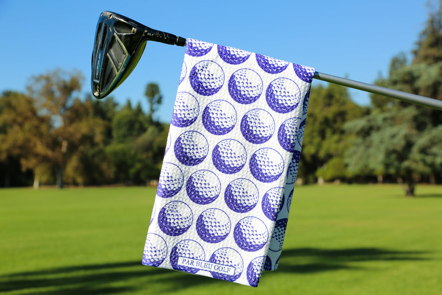 Blue golf towel with white golf ball print hanging from a club against a vibrant fairway background—perfect for adding personality to your golf bag.
