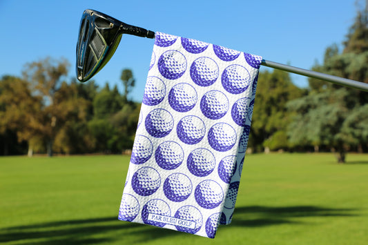Blue golf towel with white golf ball print hanging from a club against a vibrant fairway background—perfect for adding personality to your golf bag.