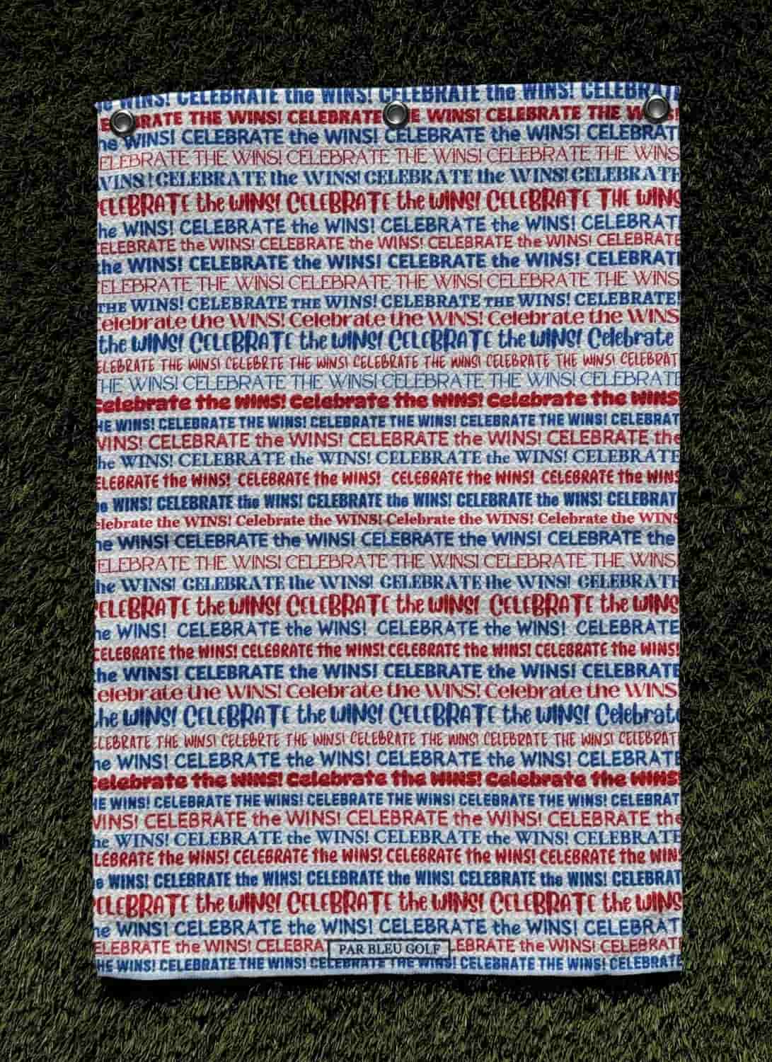 Flat red, white, and blue microfiber golf towel with “Celebrate the Wins” message, shown on turf