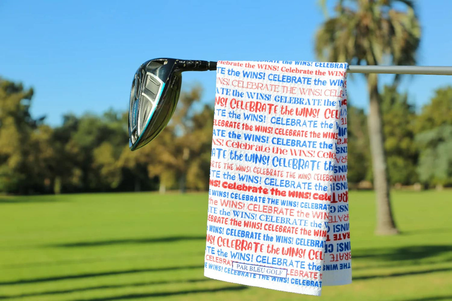 Red, white, and blue “Celebrate the Wins” golf towel hanging on club with green fairway in background
