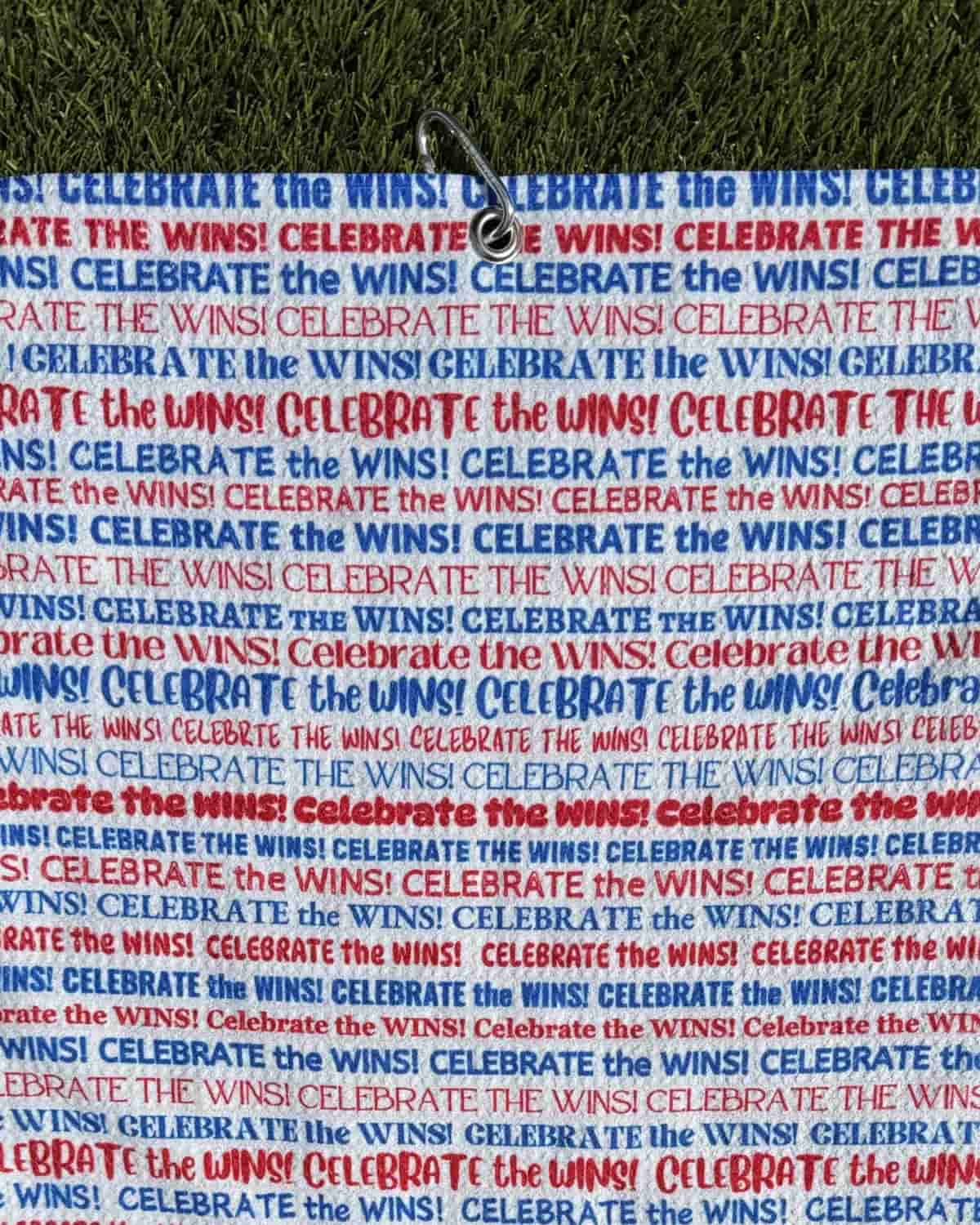 Motivational golf towel featuring “Celebrate the Wins” text in patriotic colors, shown hanging by carabiner