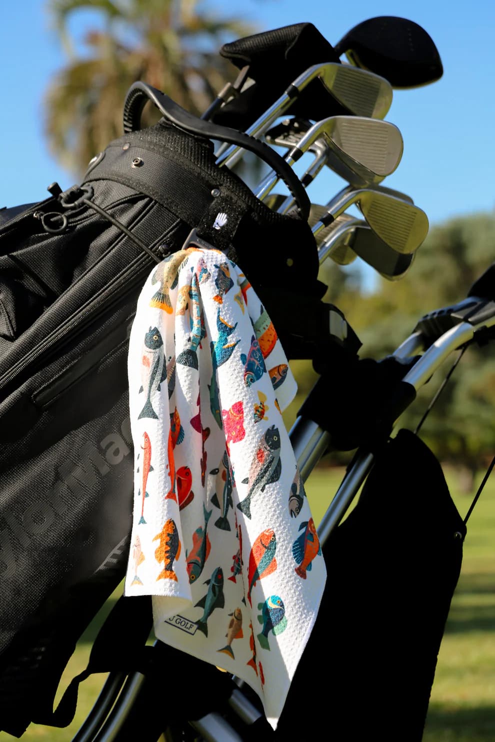 Colorful golf towel with fish pattern clipped to golf bag under sunny sky, great golf gift idea