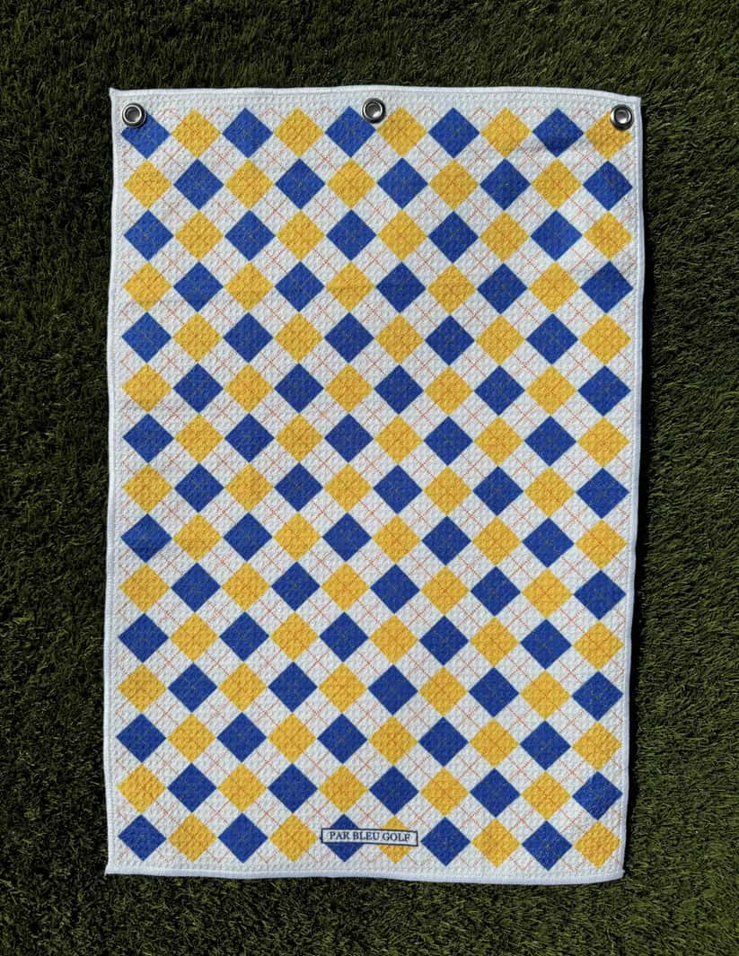 Blue and gold argyle golf towel laid flat on green grass, showcasing soft microfiber material