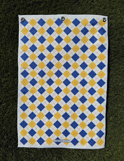 Blue and gold argyle golf towel laid flat on green grass, showcasing soft microfiber material