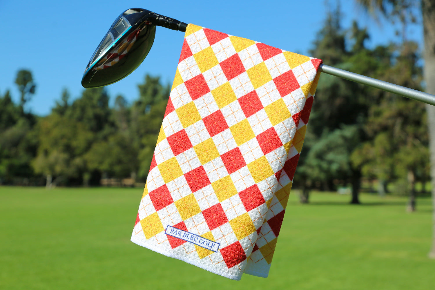 Red and gold argyle golf towel hanging on club with green grass and trees in background