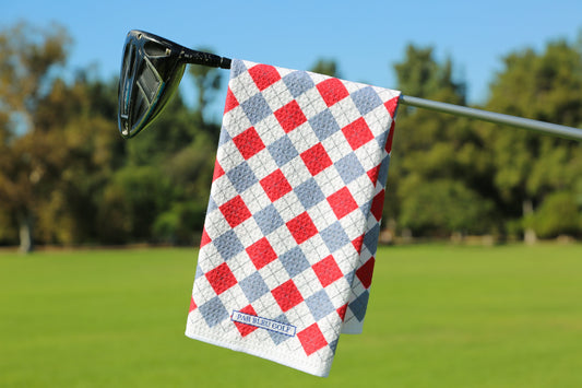 Red and grey argyle golf towel hanging on club with green fairway and blue sky in background