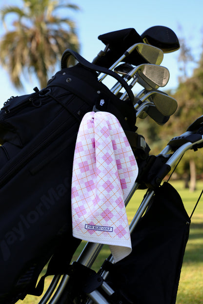Light pink argyle golf towel clipped to black golf bag with palm trees in the background