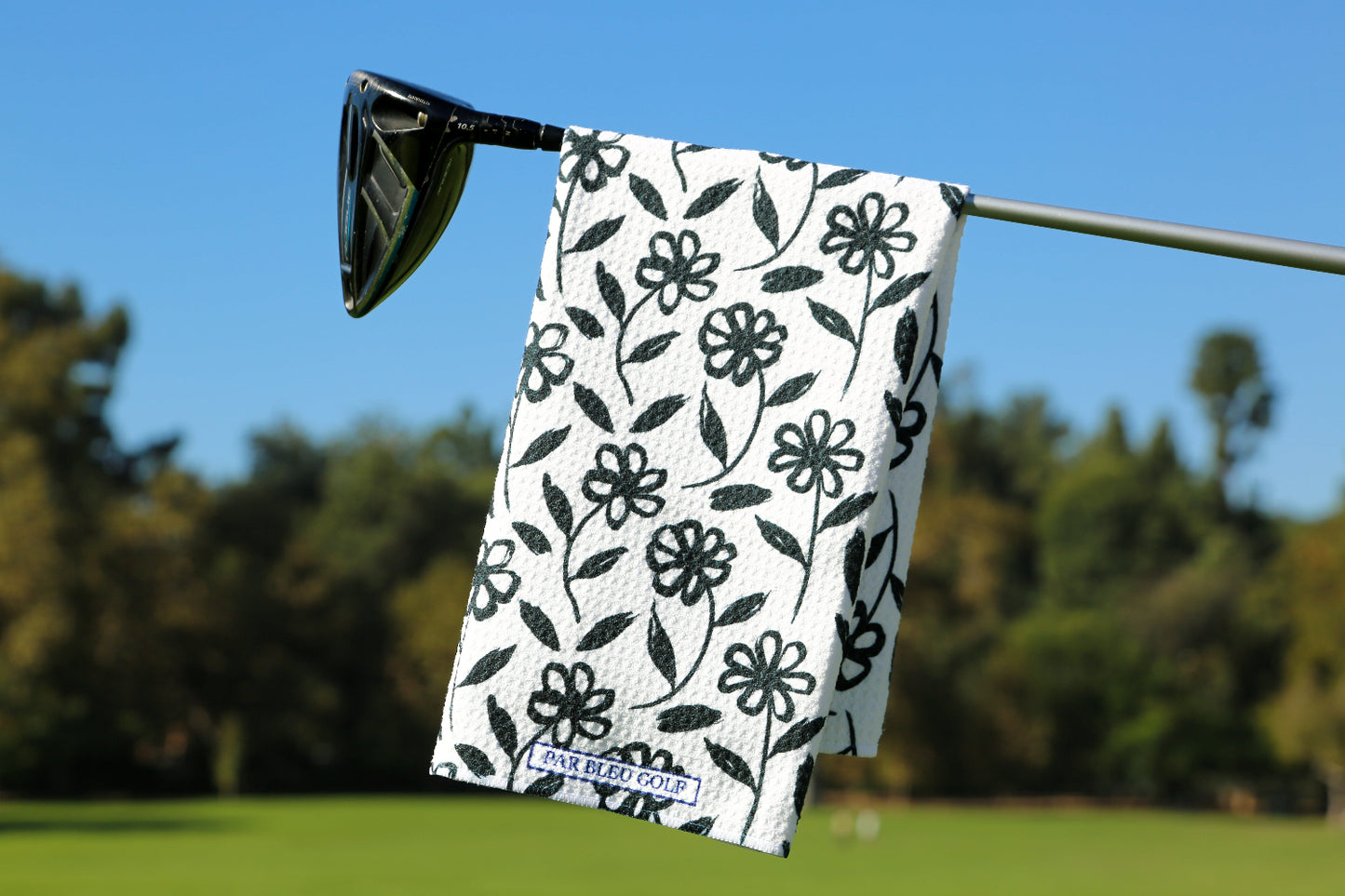 The Black Daisy golf towel hanging on a club with bold black floral print on green fairway