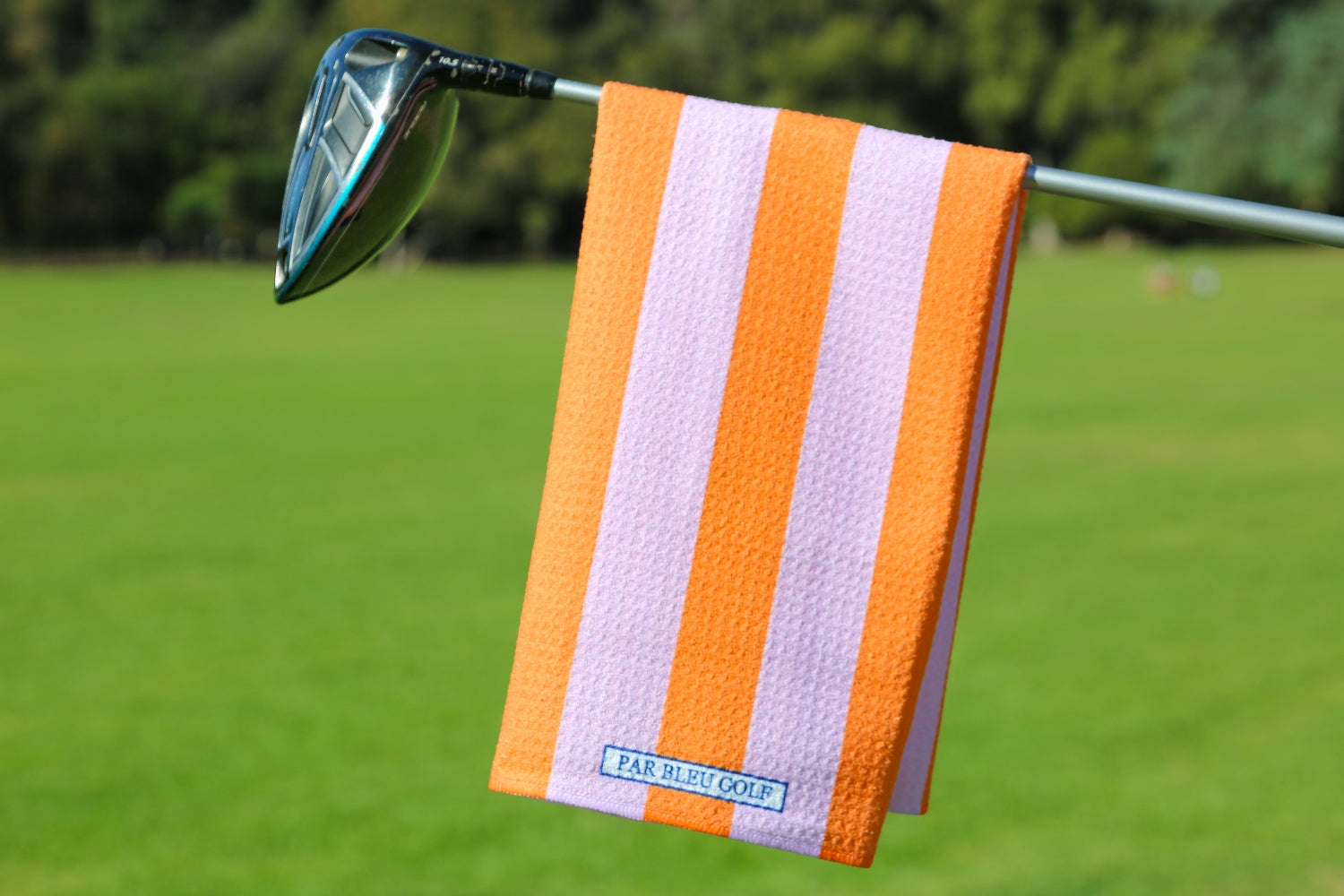 Pink and orange striped golf towel hanging on club with bright green fairway and clear sky