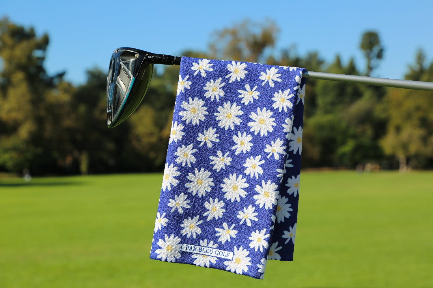 Women’s navy golf towel with white daisy floral pattern hanging on a club with green course background