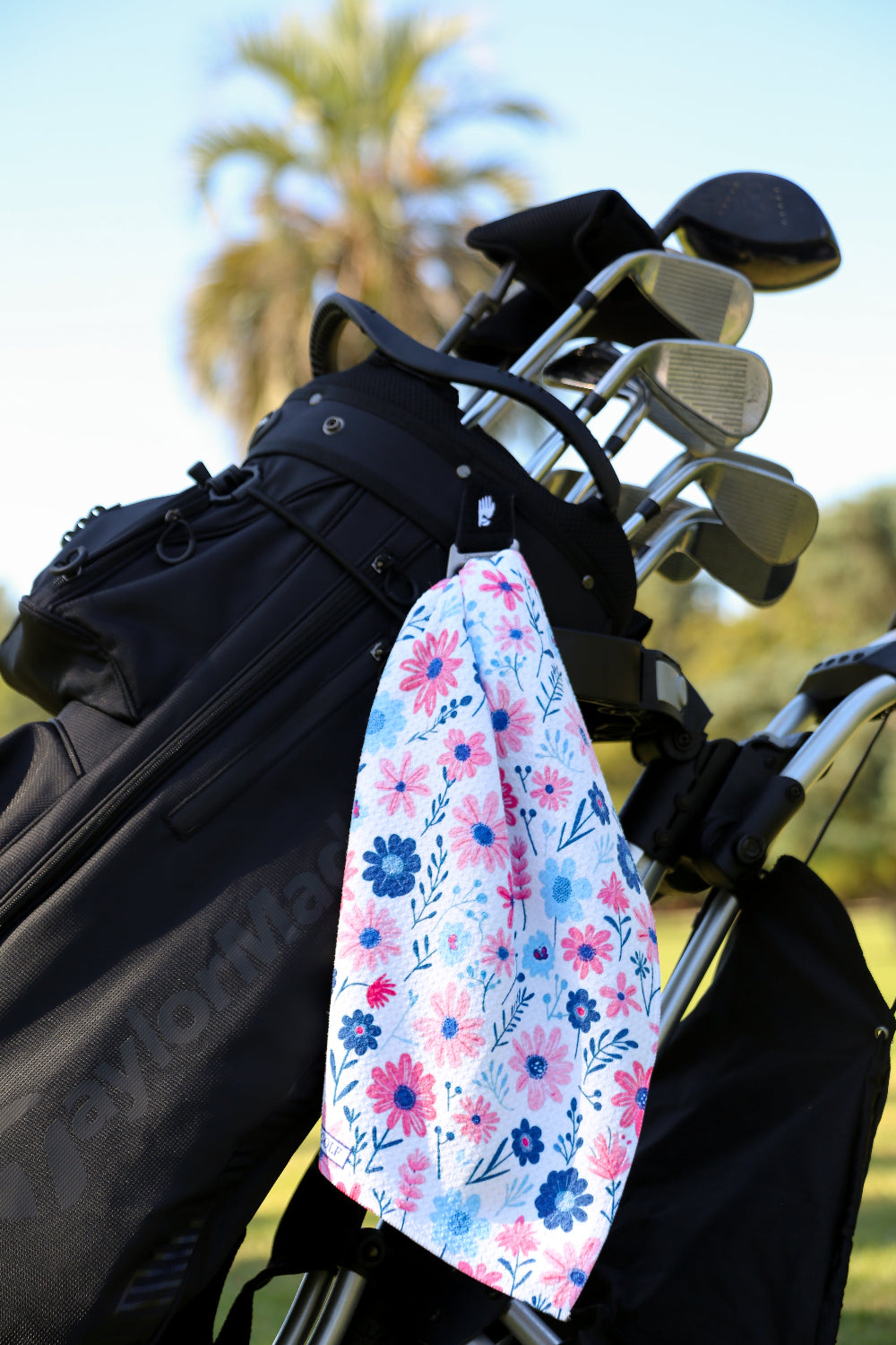 Stylish women’s floral towel clipped to black golf bag on sunny course, featuring pink and navy flower design