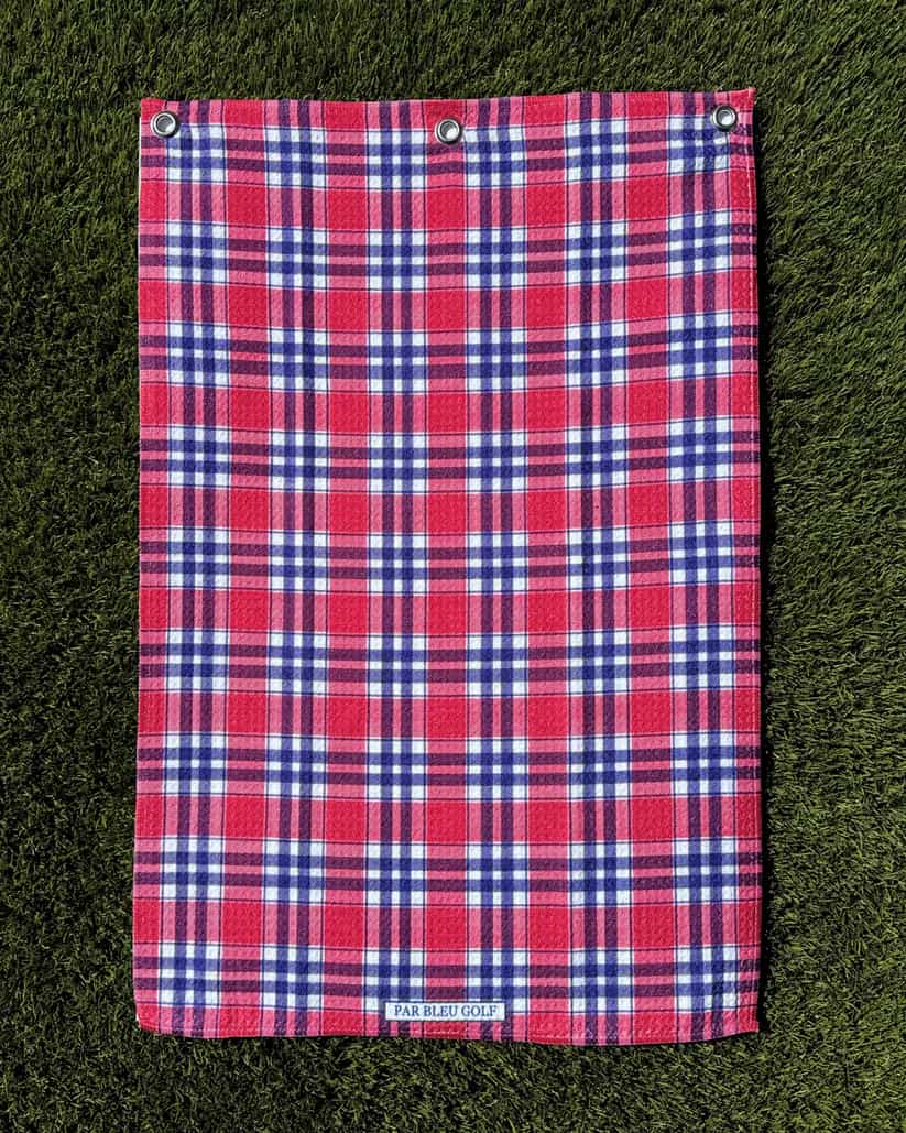 Red, navy, and white plaid towel laid flat on green grass, highlighting texture and full design