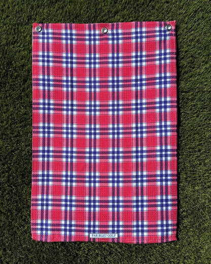 Red, navy, and white plaid towel laid flat on green grass, highlighting texture and full design