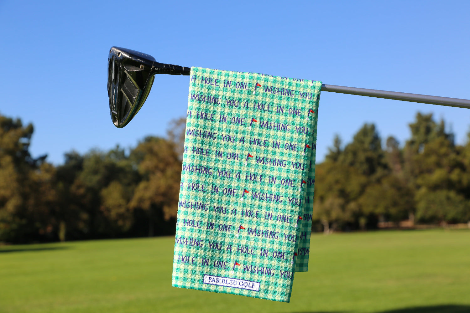 Green golf towel with “Wishing You a Hole in One” text hanging on club over scenic course background